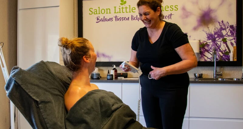 Caroline Voorhoeve lacht naar een klant in de behandelstoel, met het logo van Salon Little Wellness op de muur